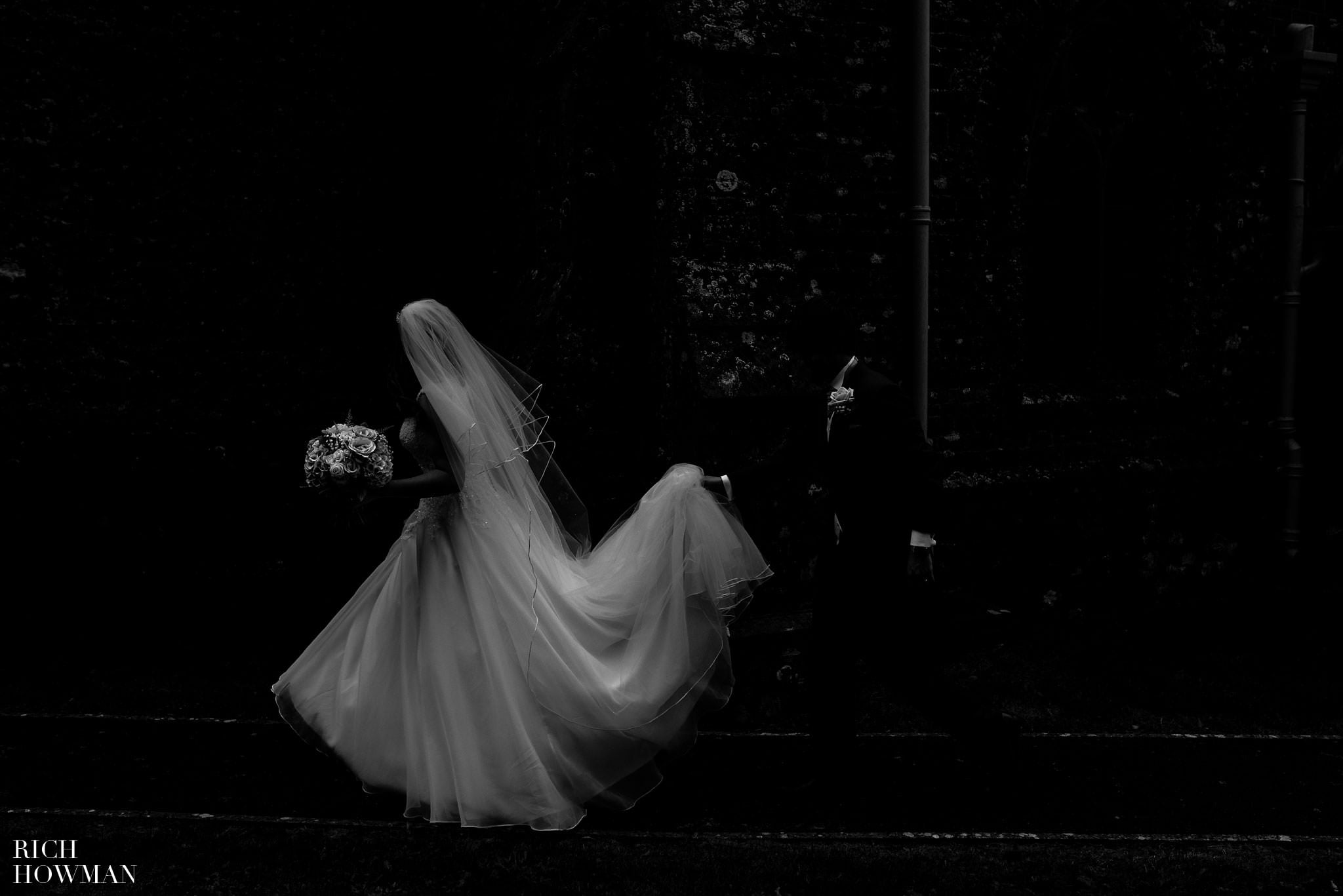 Creative black and white wedding photograph of the bride walking past the church with the groom helping her hold her wedding dress