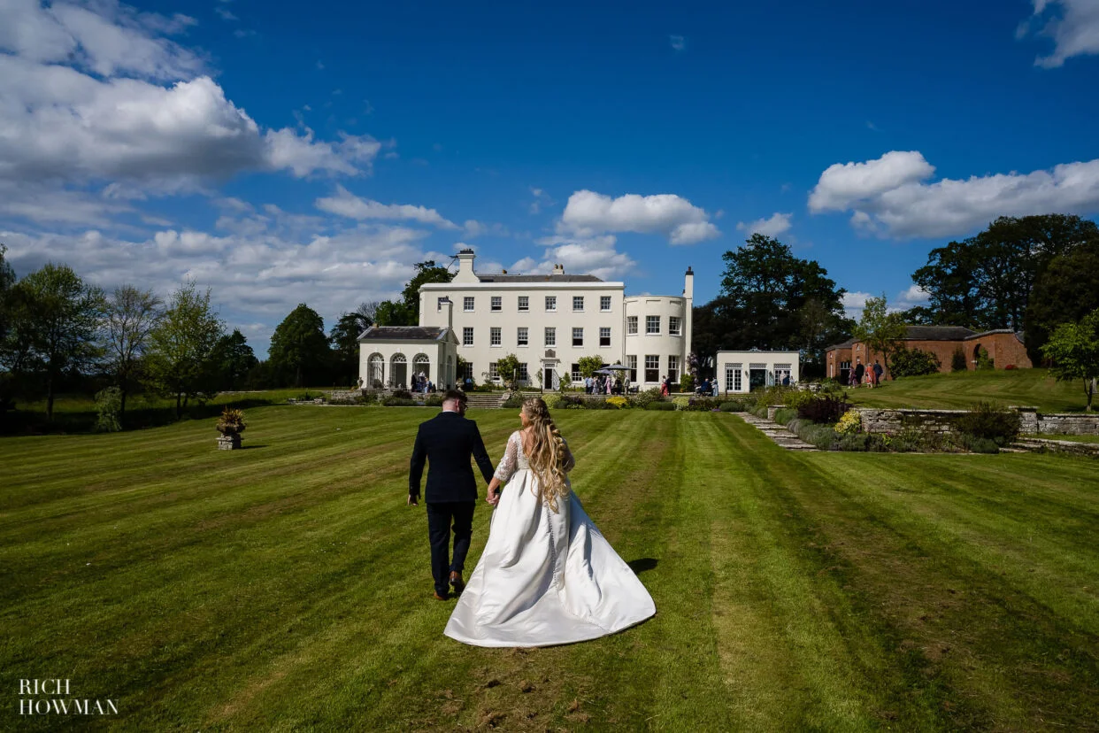 Bride and groom walking towards manor house on lawn by rockbeare manor wedding photographer, rich howman