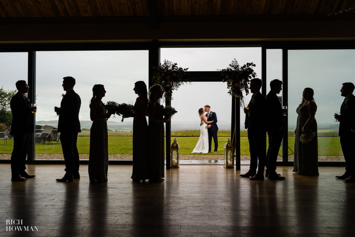 creative wedding party portrait of bride and groom kissing behind silhouettes of bridesmaids and groomsmen, by chalk barn pewsey wedding photographer, rich howman