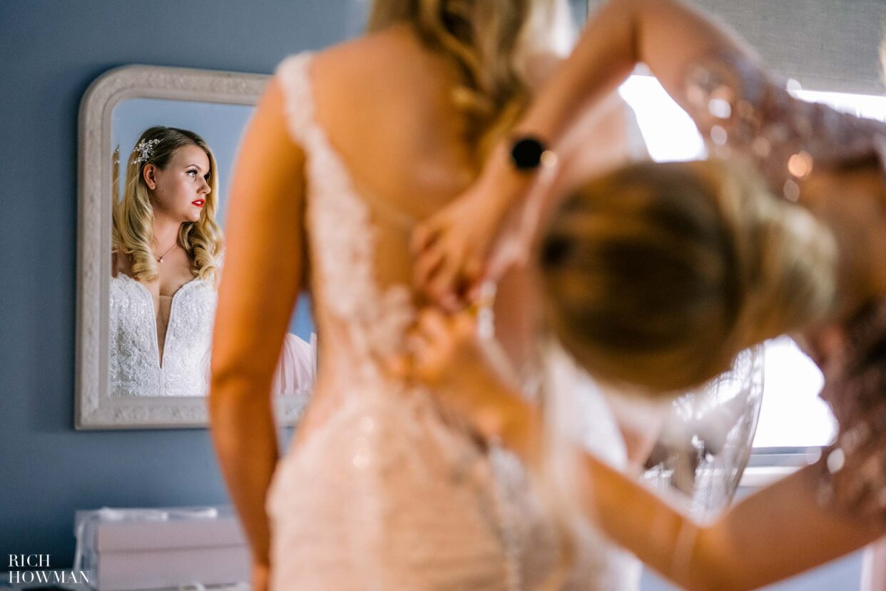bridesmaid buttoning bride's dress in front of the mirror, captured by deek park wedding photographer, Rich howman