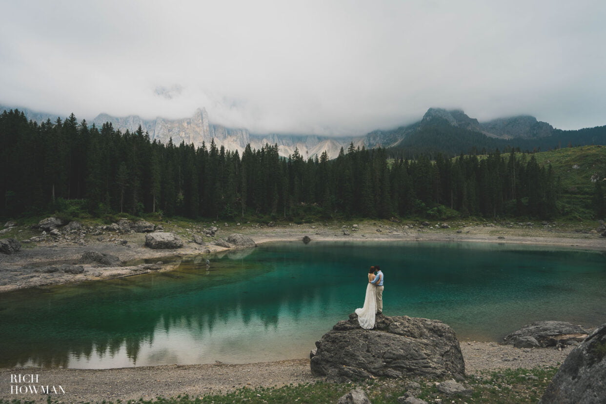 couple stand on a rock overlooking an azure lake, captured by dolomites wedding photographer, Rich Howman