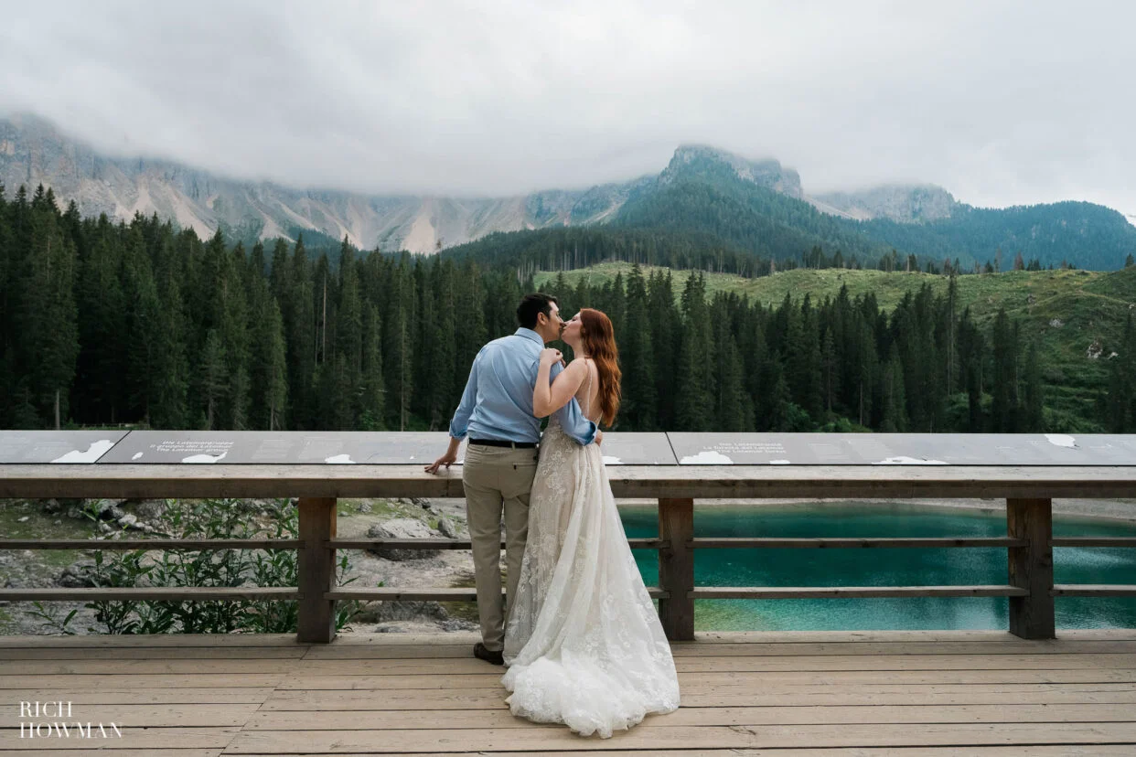 bride and groom kiss next to a lake as cloud gather over the forest, captured by dolomites wedding photographer, Rich Howman