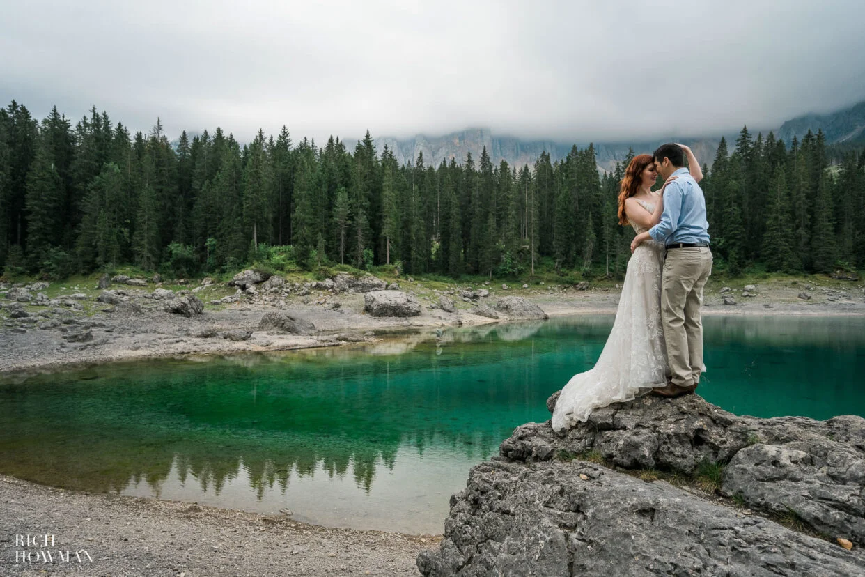 couple stnading on a rock next to vibrant azure lake and forest, captured by dolomites wedding photographer, Rich Howman