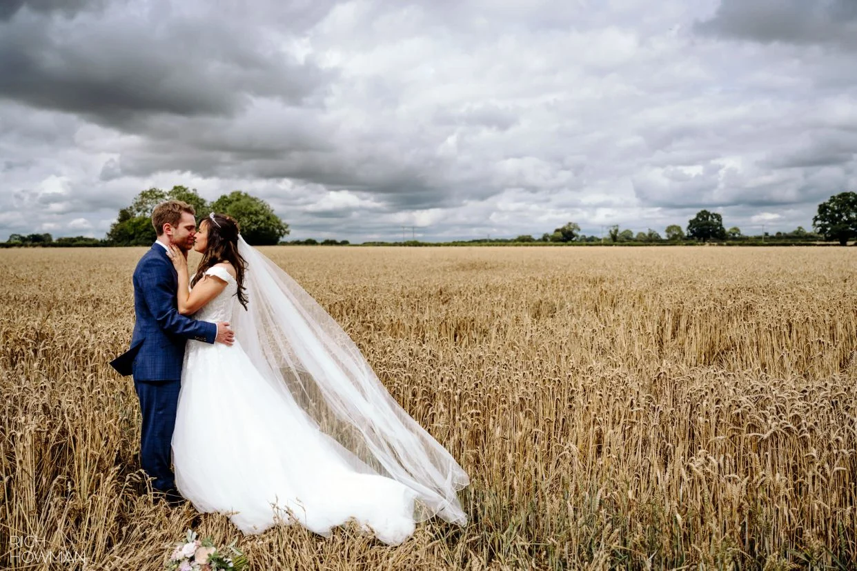 bride and groom kiss in a field with dramatic skyline, captured by tetbury wedding photographer, Rich Howman