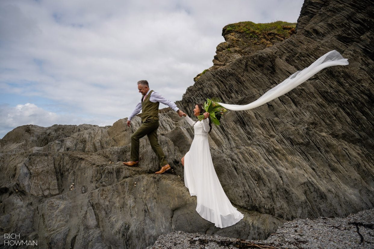 bride and groom climbing rocks at the beach with veil blowing in the wind, captured by North Devon wedding photographer, Rich Howman